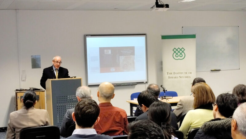 A person addressing the audience with a presentation on a projector in the back ground