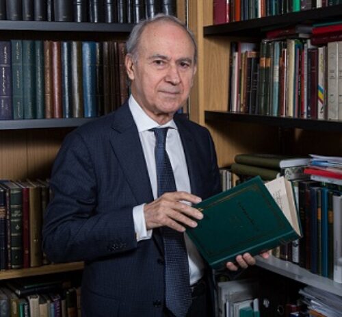 A man in suit with a book in his hand sitting in a library corner with a stack of books in the background