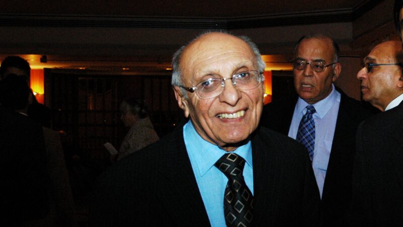 Mr Pyarali Abji Jiwa in a suit smiling and posing, with dignitaries in the back view.