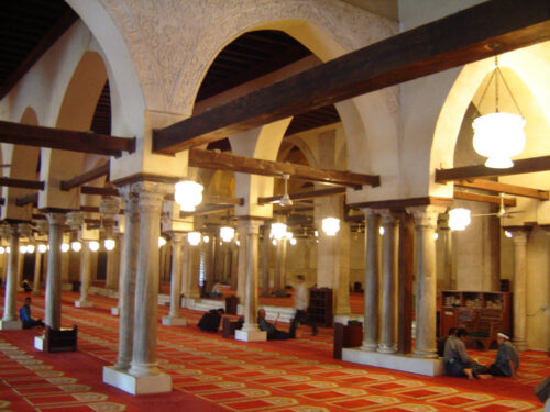 A view from inside of a prayer space with orange carpets spread through out the hall with some people sitting in different corners and praying.
