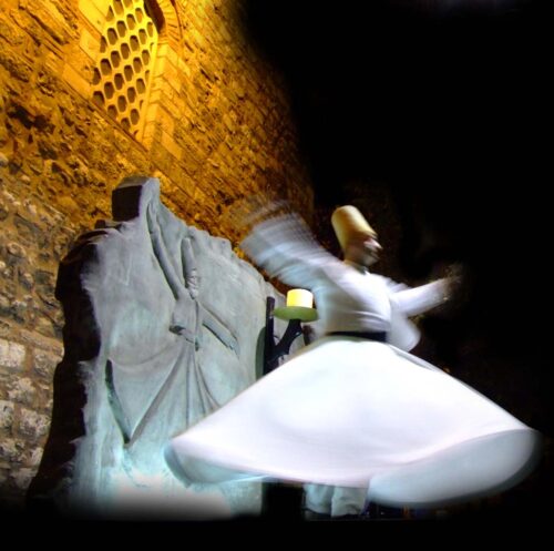 A wall with sculpted dervish carved on wood and a blurry view of a live dervish doing a Sufi dance