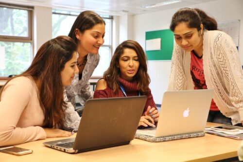 An IIS faculty leaning and observing the work in the laptops with two students sitting and one student standing and listening from the side