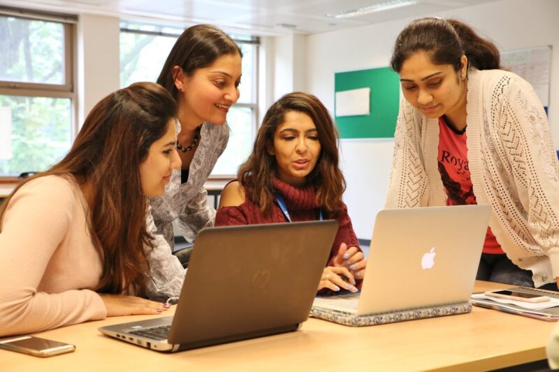 An IIS faculty leaning and observing the work in the laptops with two students sitting and one student standing and listening from the side