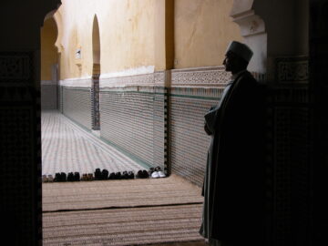 A man dressed in traditional clothes patiently standing in the corner of a prayer hall and praying