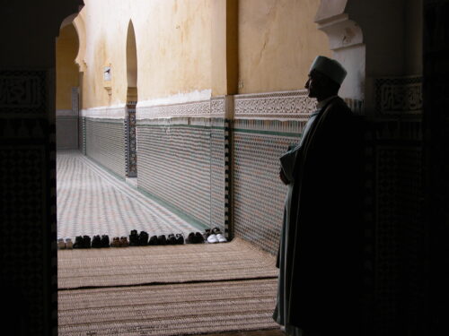 A man dressed in traditional clothes patiently standing in the corner of a prayer hall and praying