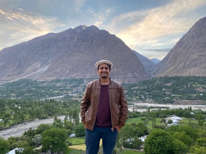A man in brown leather jacket and beret standing in a hilly area of Chitral Pakistan smiling and posing