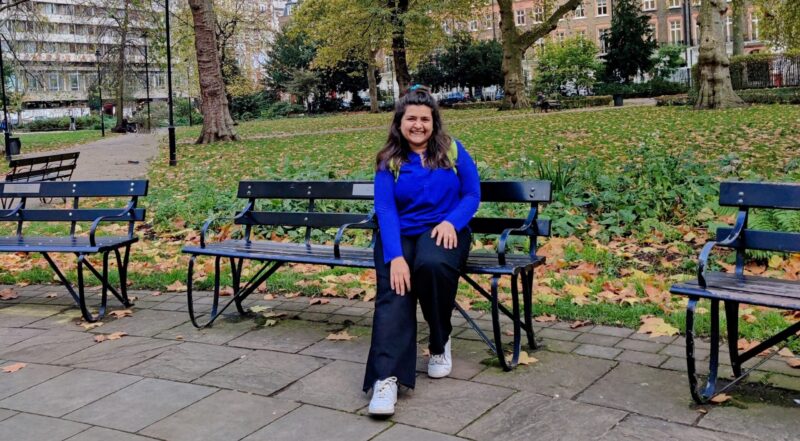 A young lady sat on a bench in a London square with grass and fallen leaves behind her