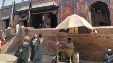 Three Muslim women standing outside of the shrine of Pir Shams near a man sitting under an umbrella selling rose petals.