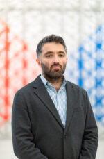 Aslisho Qurboniev in a grey blazer and a blue shirt posing for a picture.