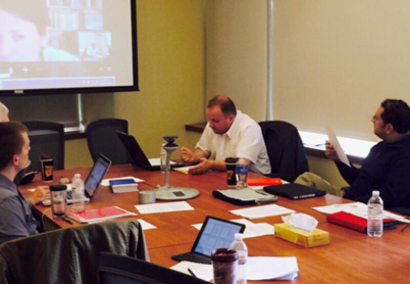 A group of people sitting over a round table and having an in-person as well as virtual meeting via online platform