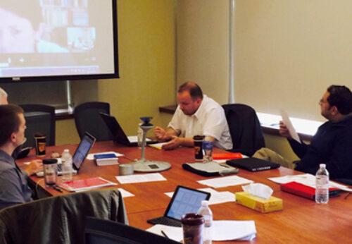 A group of people sitting over a round table and having an in-person as well as virtual meeting via online platform