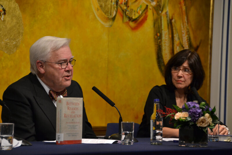 A man in suit sitting and speaking over a podium and a woman next to him looking at him