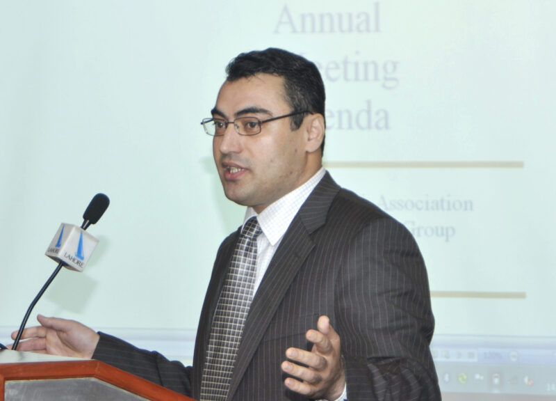 A side view of a man in suit speaking over a podium with a presentation in the background