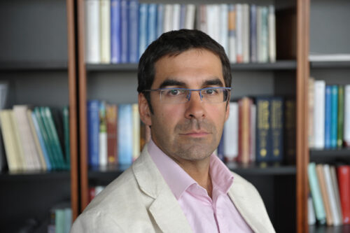 A man in white blazer , pink shirt, and a stack of books behind him