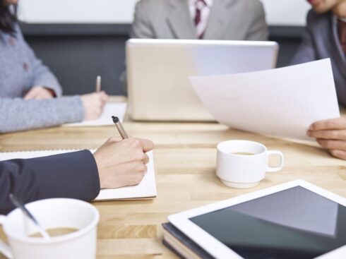 Two coffee mugs, one tablet and a few open laptops with some stationery items lying on the table and some hands of people to reflect an active working space