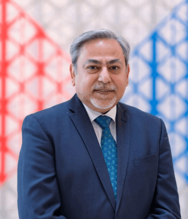 A well-dressed man in a blue suit and patterned tie stands before a colourful geometric-patterned background.