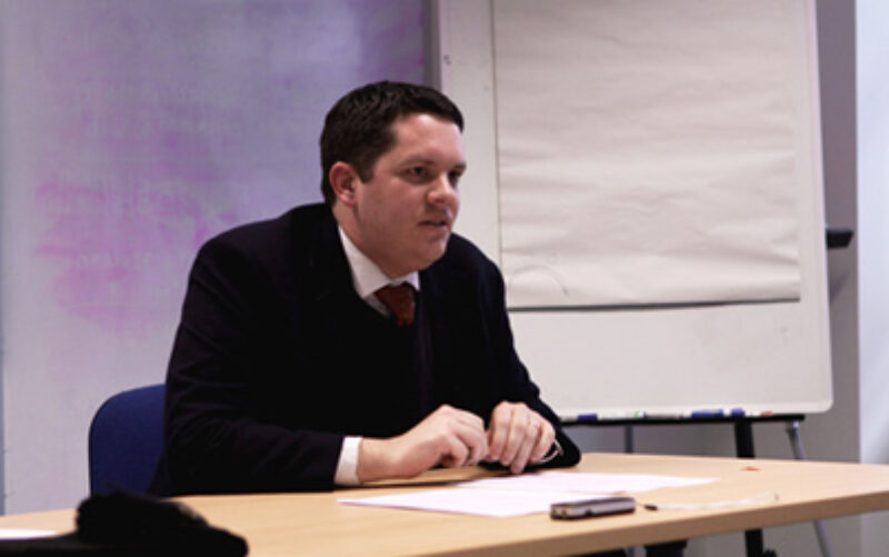 A man in black suit sitting and addressing a audience