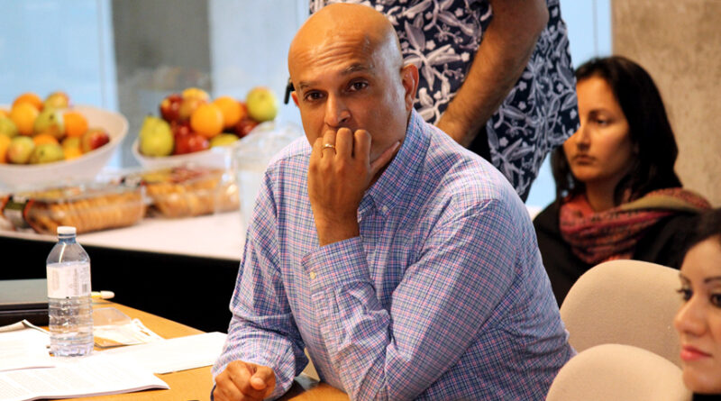 A man in blue shirt sitting on a table with an empty bottle wandering with fingers on his mouth, with some baskets of fruits and people in the blurry background